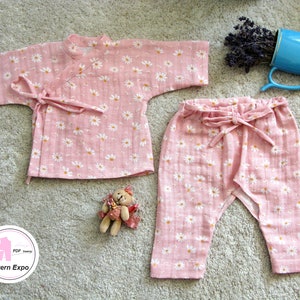Könnte beinhalten: Ein rosa Baby-Outfit mit einem Gänseblümchen-Druck. Das Oberteil ist im Kimono-Stil mit einem Bindeverschluss. Die Hose hat einen elastischen Bund und eine Schleife an der Taille.