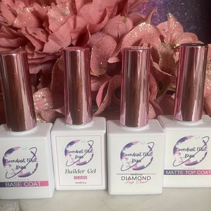 May include: Four bottles of nail polish with pink lids and white labels. The labels have a logo with a planet and the text "Goondust Nail Dips". The bottles are labeled "Base Coat", "Builder Gel Clear 15ml/0.5 fl oz", "Diamond Top Coat", and "Matte Top Coat".