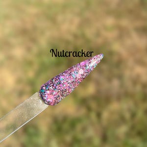May include: A close-up of a nail polish color called "Nutcracker". The polish is a mix of pink, purple, and teal glitter.