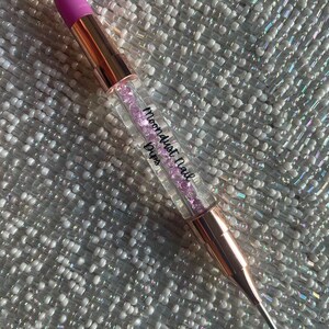 May include: A rose gold nail art tool with a purple silicone tip and a clear body filled with pink glitter. The tool is labelled "Moondust Nail Drips".