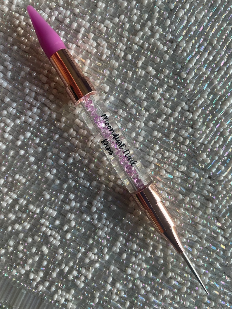 May include: A rose gold nail art tool with a purple silicone tip and a clear body filled with pink glitter. The tool is labelled "Moondust Nail Drips".
