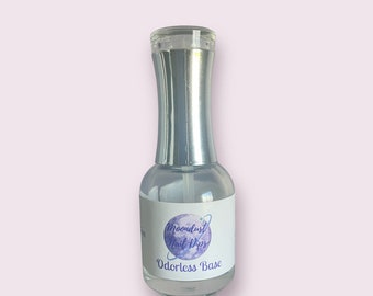 May include: A clear glass bottle of nail dip base with a silver cap. The label features the text "Moondust Nail Dips" and "Odorless Base" in a purple and white design. The bottle is on a light pink background.