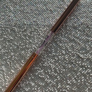 May include: A rose gold makeup brush with a clear handle filled with pink glitter. The handle has the text "Glamdust Trail" and "POPPY" printed on it.