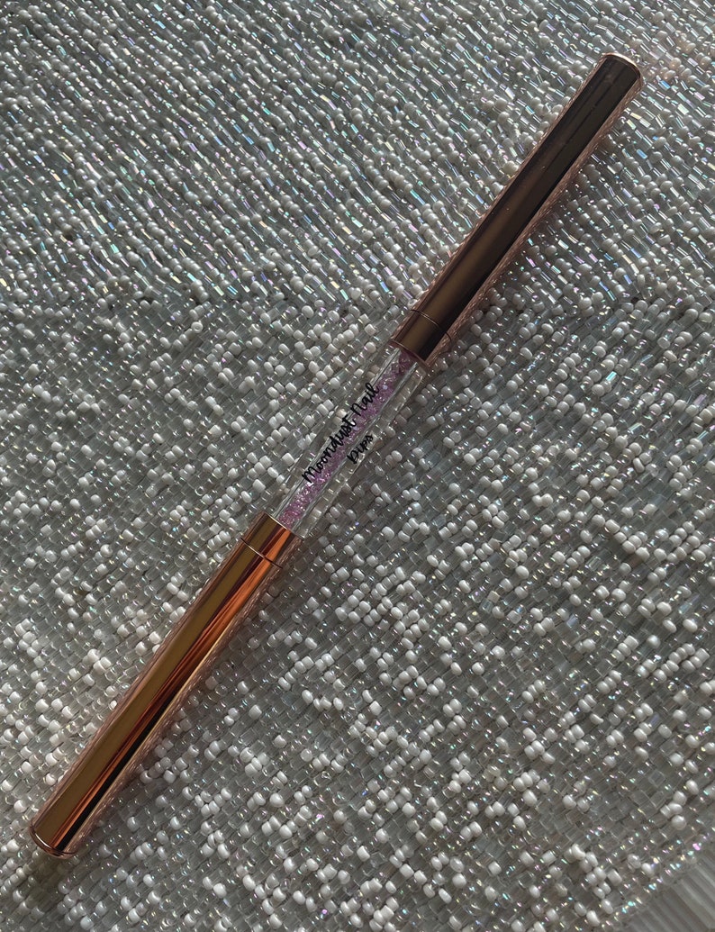 May include: A rose gold makeup brush with a clear handle filled with pink glitter. The handle has the text "Glamdust Trail" and "POPPY" printed on it.