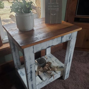 May include: A whitewashed wooden end table with a distressed finish. The table has a top shelf and a lower shelf. A small potted plant and a wooden sign with the text "do not let your heart be troubled" are on the top shelf.
