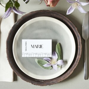 May include: A white place card with the text "WELCOME" and "MARIE" printed on it. The place card is on a brown plate with a grey plate underneath. There are green leaves and a white flower on the plate.