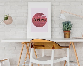 May include: A white framed print with a pink background and a red circle with the word "Aries" in black text. The print is hanging on a white brick wall above a white desk with a wooden chair and a potted plant.