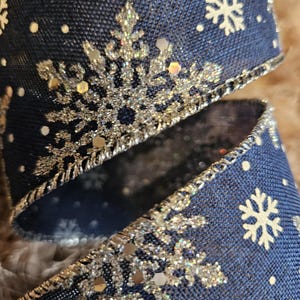 Könnte beinhalten: Nahaufnahme eines marineblauen Bandes mit Schneeflockenmuster. Das Band hat silberne Glitzer-Schneeflocken und weiße Punkte. Die Kanten sind mit silbernem Metallic-Garn verziert, was einen festlichen und dekorativen Look erzeugt.