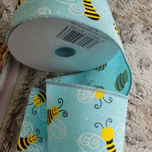 Op de afbeelding: Een rol blauw lint met een wit en geel bijenpatroon. Het lint is 2,5 inch breed en heeft een bedrade rand. Het etiket op de rol luidt "960232 021133  2,5" wired edge skinny  NO. 30F  bee"