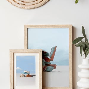 May include: Two framed art prints with light wood frames. The prints depict stacked stones against a blue sky and white background. A woven wall hanging and a white vase with greenery are also visible.