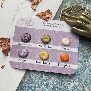 May include: A watercolour sample set named "Enchanted Cauldron" with six colours: Enchanted Plum, Witch's Bog, Bones, Potion, Star Light, and Pumpkin. The colours are on a purple card with the brand name "Pano Phobia".