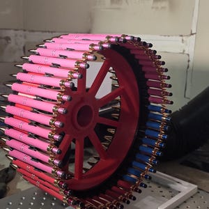 May include: A circular display of pens in shades of pink, red, and blue, arranged around a red wheel. The pens have gold accents. A purple notebook with text is in the foreground.