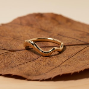 Könnte beinhalten: Ein goldfarbener Ring mit wellenförmigem Design. Der Ring liegt auf einem braunen Blatt, was einen Kontrast aus Texturen und Farben erzeugt. Die glatten, geschwungenen Linien des Rings verleihen ihm ein modernes und elegantes Aussehen. Ein Schmuckstück.