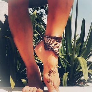 Può includere: Un braccialetto alla caviglia marrone e bianco con chiusura a bottone è indossato su un piede con un tatuaggio all'henné. Lo sfondo comprende piante verdi e un muro bianco.