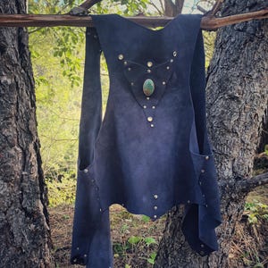 Puede incluir: Un chaleco de ante azul oscuro con un panel trasero triangular adornado con una piedra verde ovalada central y pequeños remaches metálicos. La prenda tiene un diseño abierto con paneles laterales sujetos por remaches plateados.