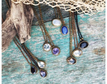 May include: A collection of necklaces with various gemstone pendants. The pendants are oval and teardrop shaped and feature a variety of colors, including clear, purple, blue, black, and brown. The necklaces are made with brown cord and have a rustic, bohemian aesthetic.