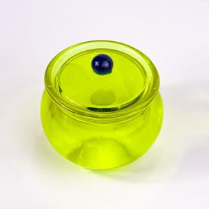 Könnte beinhalten: Ein kleines, rundes, limettengrünes Glasgefäß mit passendem Deckel. Der Deckel ist mit einer kleinen, dunkelblauen Glaskugel versehen. Das Gefäß ist transparent, so dass Licht hindurchscheinen kann. Das Gefäß wird wahrscheinlich zur Aufbewahrung oder Dekoration verwendet.