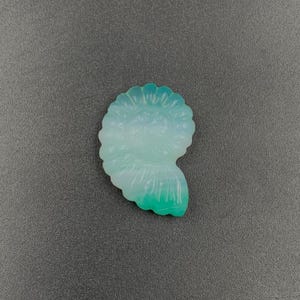 May include: A translucent, seashell-shaped carving in shades of light blue and green. The polished surface reflects light, highlighting the intricate spiral design. The object is set against a gray background.