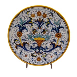 Puede incluir: Un plato redondo de cerámica con un diseño pintado a mano y un borde amarillo. El centro muestra un motivo de cesta con frutas amarillas, rodeado de intrincados adornos azules, hojas verdes y flores naranjas sobre un fondo blanco.