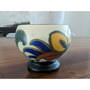 May include: A white ceramic bowl with a black base and a colorful floral design. The design features yellow, orange, blue, and green colors.