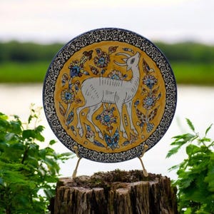 May include: A round ceramic plate with a yellow background and a white deer with blue and brown floral designs. The plate is on a wooden stand.