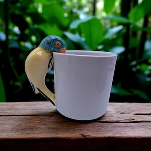 Puede incluir: Taza de cerámica blanca con un asa en forma de pájaro. El asa es una escultura detallada de un pájaro con colores azul, amarillo y naranja. La taza está colocada sobre un fondo verde borroso, lo que sugiere un entorno al aire libre.