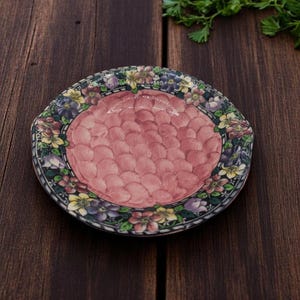 Op de afbeelding: Een decoratieve keramische schaal met een roze midden en een bloemenrand. De schaal heeft een geschulpte rand en is versierd met een verscheidenheid aan kleurrijke bloemen, waaronder geel, paars en wit. De schaal staat tegen een donkere houten achtergrond.