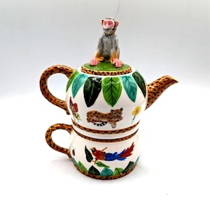 Könnte beinhalten: Dekoratives Tee-Set mit einer Affenfigur auf dem Deckel. Das weiße Keramik-Set ist mit grünen Blättern, einem Leoparden, einem Schmetterling und einem Papagei verziert. Die Griffe und der Ausguss haben ein braunes Leopardenmuster.