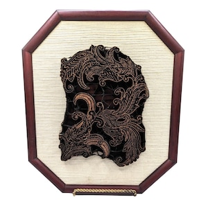 Pode incluir: Uma peça de arte de parede batik enquadrada com uma moldura de madeira marrom e um fundo bege. O design batik é marrom escuro e apresenta um padrão floral.