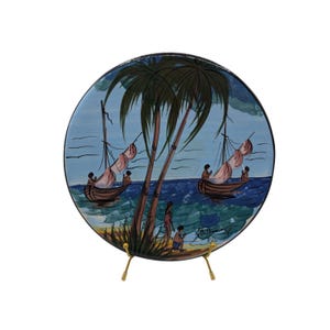 May include: A round decorative plate with a hand-painted tropical scene. The plate features sailboats on the ocean, palm trees, and figures on the shore. The color palette includes blue, brown, green, and pink.