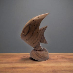 Könnte beinhalten: Holzfischskulptur mit geschwungener Flosse und Schwanz sowie einem abgerundeten Sockel. Der Fisch ist mittelbraun mit kleinen, dunklen Punkten und einem goldfarbenen Auge. Die Skulptur steht auf einer Holzoberfläche.