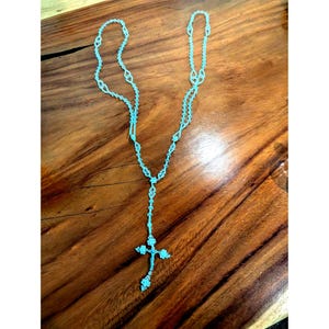 May include: A light blue rosary necklace with a cross pendant. The necklace is made of small, connected beads and is displayed on a wooden surface. The cross pendant is also light blue and has a detailed design.