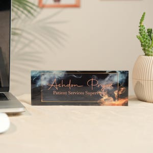 May include: A rectangular desk nameplate with the name "Ashdon Pryor" and the title "Patient Services Supervisor" in a copper-colored script. The nameplate has a black background with a smoky design and a gold border. A laptop and a potted plant are in the background.