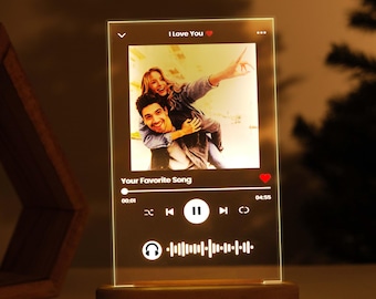 Op de afbeelding: Een heldere acryl lamp met een foto van een stel en een muziekspelerinterface. De lamp heeft een houten voet en een rood hartpictogram. De tekst op de lamp luidt "Ik hou van je" en "Je favoriete nummer".