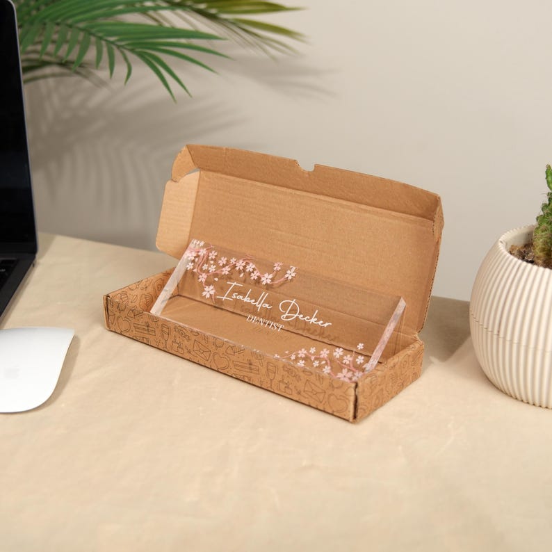 May include: A clear acrylic desk nameplate with the name "Isabella Decker" and the title "Dentist" in white script, adorned with pink floral accents. The nameplate is displayed inside an open cardboard box.