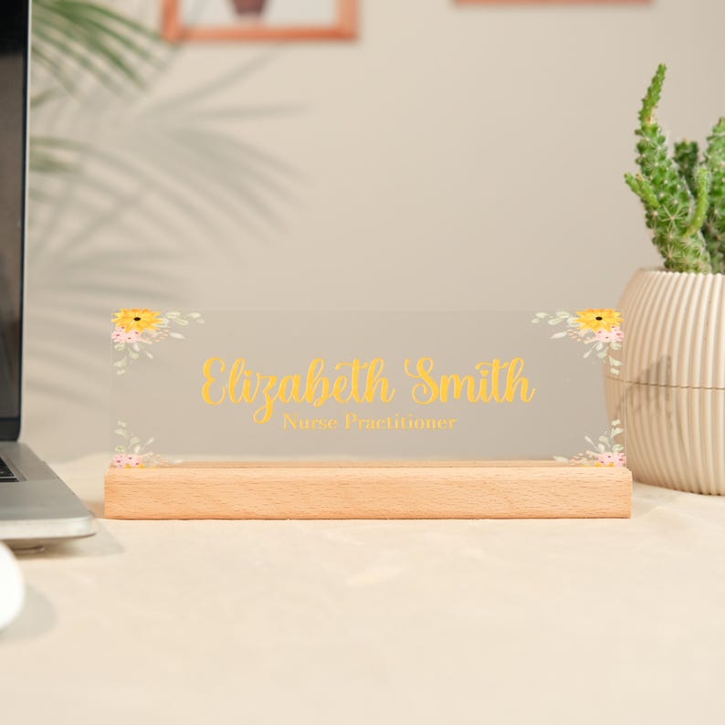 May include: A clear acrylic desk nameplate with the name "Elizabeth Smith" in gold script, and the title "Nurse Practitioner". The nameplate is decorated with floral accents and sits on a wooden base.