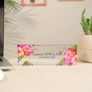 May include: A clear acrylic desk nameplate with floral accents. The nameplate features the name "Aurora SINCLAIR" and the title "PHARMACIST" in black text. Pink, red, and green floral designs adorn the corners.