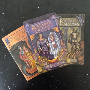 May include: Three fantasy paperback books by Deborah Chester titled Shadow War, Realm of Light, and Reign of Shadows. Each cover features detailed illustrations of figures in medieval-style clothing, swords, and dragons with purple and gold tones.