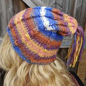 May include: A hand-knitted beanie featuring a variegated yarn pattern in shades of blue, orange, yellow, and purple. The hat has a textured stitch design, a prominent white vertical stripe, and a long, multi-coloured yarn tassel attached to the side.