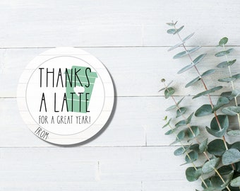 May include: Round white sticker with black text that says "Thanks a latte for a great year!" with a green coffee cup icon. The sticker is on a white wood background with green eucalyptus branches.