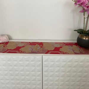 May include: A white cabinet with two doors and a red and gold patterned table runner on top. A pink orchid plant in a black pot is on the right side of the table runner.