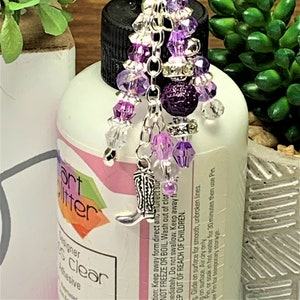 May include: A clear bottle with a purple and silver charm hanging from the lid. The charm features a purple and silver boot, purple and clear beads, and a sparkly purple ball. The bottle is labeled "Art Glitter Designer Glue Clear Adhesive".