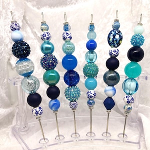 May include: A collection of seven beaded skewers in shades of blue, teal, and white. Each skewer features a variety of bead shapes and textures, including round, faceted, and patterned beads. The skewers are displayed in a clear plastic stand.