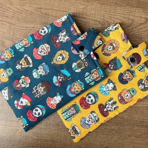Op de afbeelding: Twee stoffen tasjes met een ritssluiting en een drukknoop. De tasjes zijn versierd met een herhalend patroon van cartoondieren die zonnebrillen en hoeden dragen. Eén tasje is blauw en de andere is geel.