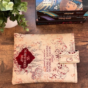 Può includere: Una copertina per libri in tessuto beige con un disegno a mappa con accenti rossi e la scritta "The Marauders Map". Ha una chiusura a patta abbinata. Diversi libri di "Harry Potter" sono impilati sullo sfondo.