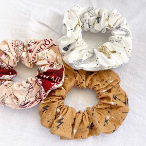 Può includere: Tre scrunchies in tessuto con diverse stampe a tema Harry Potter. Uno scrunchie è bianco con stelle dorate e un boccino d'oro, uno è beige con accenti rossi e marroni e una stampa di mappa, e l'ultimo è marrone con stelle dorate e fulmini.