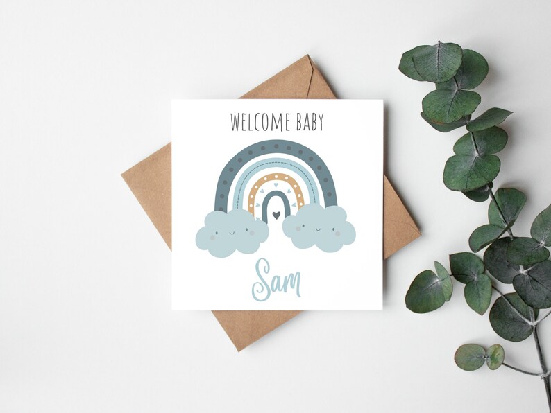 Square card with blue and gold boho-style rainbow. At either end of rainbow there are cute, fluffy clouds with smiley faces. The card reads Welcome Baby Sam