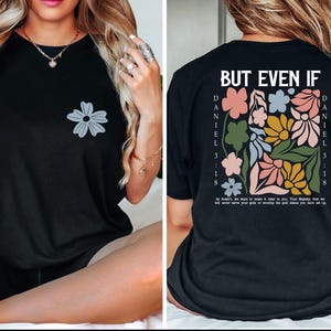 May include: Black t-shirt with a gray flower graphic on the front. The back features a floral design with the text "BUT EVEN IF" and "DANIEL 3:18". The shirt has a relaxed fit and short sleeves.