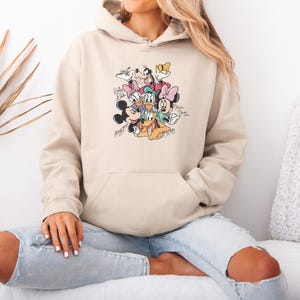 May include: Beige hooded sweatshirt featuring a colorful graphic of Disney characters including Mickey Mouse, Minnie Mouse, Donald Duck, and Goofy. The graphic includes the characters' signatures. The sweatshirt has a front pocket and a drawstring hood.