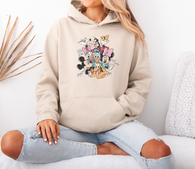 May include: Beige hooded sweatshirt featuring a colorful graphic of Disney characters including Mickey Mouse, Minnie Mouse, Donald Duck, and Goofy. The graphic includes the characters' signatures. The sweatshirt has a front pocket and a drawstring hood.
